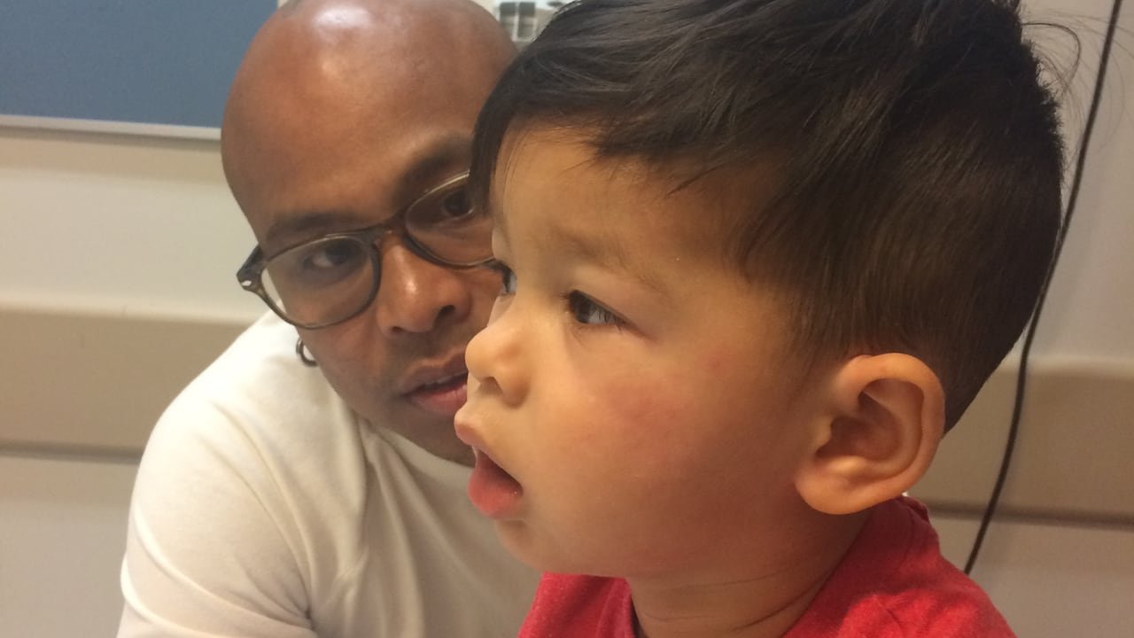 default-165 In a hospital room, a man looks concerned at a boy breathing with his mouth open and who has hives on his cheeks.